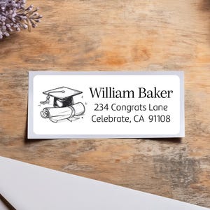 Peut inclure: Une étiquette rectangulaire blanche avec une illustration d'un chapeau de graduation et d'un diplôme. L'étiquette indique le nom "William Baker" et une adresse : 234 Congrats Lane, Celebrate, CA 91108.