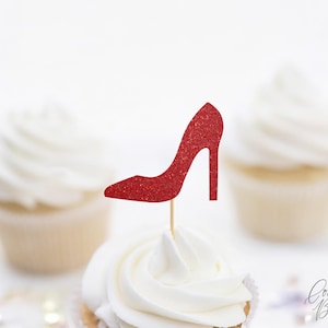 May include: A red glitter high-heeled shoe cupcake topper on a cupcake with white frosting. The cupcake is in front of two other cupcakes with white frosting. The image has a soft, bright background.
