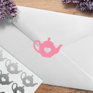 May include: A white envelope with a pink teapot sticker featuring a heart-shaped cutout. A sheet of silver teapot stickers is visible on the left. Purple flowers are in the background. The image is on a wooden surface.