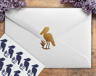 Könnte beinhalten: Ein weißer Papierumschlag mit einem metallisch goldenen Schuhschnabel-Aufkleber in der Mitte der Klappe. Daneben liegt ein Bogen mit dunkelblauen Aufklebern, die jeweils die Silhouette desselben Schuhschnabels neben hohem Gras zeigen.