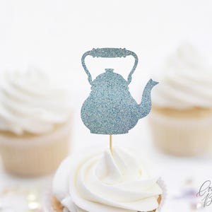 May include: A blue glitter teapot cupcake topper on a wooden stick, placed on top of a cupcake with white frosting. Two other cupcakes with white frosting are in the background. The image has a soft, blurred background.