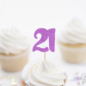 May include: A purple glitter number 21 cupcake topper featuring a stylized font attached to a thin wooden pick. The topper is positioned upright in the center of a white frosted cupcake, with two additional frosted cupcakes visible in the background.