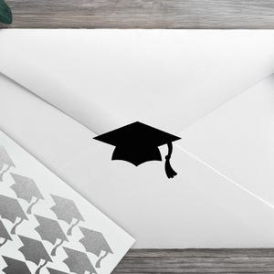 May include: A white envelope featuring a black graduation cap silhouette sticker centred on the flap, accompanied by a sheet of matching silver graduation cap stickers arranged in a grid pattern on a white rectangular backing sheet.