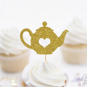 May include: A gold glitter teapot cupcake topper with a heart-shaped cutout. The topper is on a wooden stick and placed on top of a cupcake with white frosting. The background features other cupcakes.