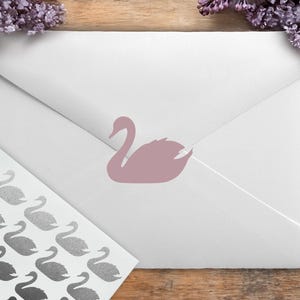 May include: A white envelope with a mauve swan sticker and a sheet of swan stickers. The stickers are in a gradient of gray. The background is a wooden surface with purple flowers.