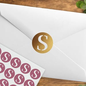 Peut inclure: Une enveloppe blanche scellée par un autocollant circulaire doré portant la lettre S dans une police serif. À côté de l'enveloppe se trouve une feuille d'autocollants circulaires rose poudré, chacun imprimé avec la même lettre S blanche.
