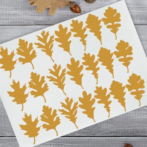 May include: A sheet of white paper with multiple golden oak leaf cutouts. The leaves are arranged in a scattered pattern. The image also includes real oak leaves and acorns, suggesting a fall theme. The logo "Confetti Barn" is in the upper left corner.