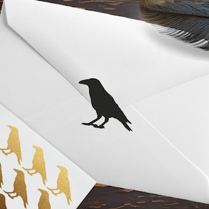 May include: A white envelope featuring a black raven silhouette on the flap, accompanied by a matching white card decorated with a repeating pattern of metallic gold raven silhouettes, positioned near a dark feather and a wooden pen.