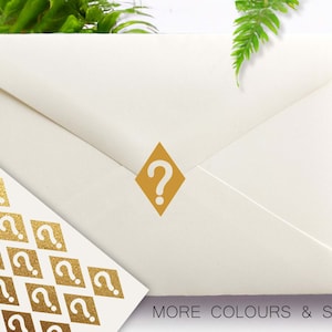 May include: A white envelope with a gold diamond-shaped sticker featuring a question mark. The envelope is surrounded by green foliage. The text "MORE COLOURS & SIZES" is printed at the bottom of the envelope.