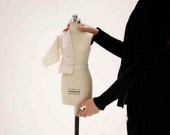 May include: A white fabric garment is being fitted on a half-scale dress form. The dress form is on a black stand. The person fitting the garment is wearing a black jacket.