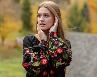 May include: A woman with long blonde hair is wearing a black embroidered blouse with red, green, and purple floral patterns. She is standing in front of a blurred background of fall foliage.
