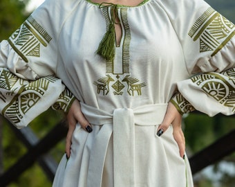 May include: A white linen dress with green embroidered geometric patterns and a green tassel at the neckline. The dress has a belt and long sleeves.