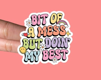 May include: A colorful sticker with the text "Bit of a mess but doin' my best" in a retro font. The sticker features flowers, a rainbow, a heart, and a smiley face.