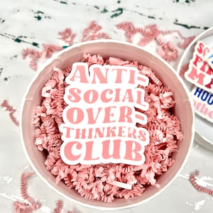 May include: A pink and white sticker with the text "Anti-Social Over-Thinkers Club" in a retro font style. The sticker is in a white round container filled with pink crinkle paper.