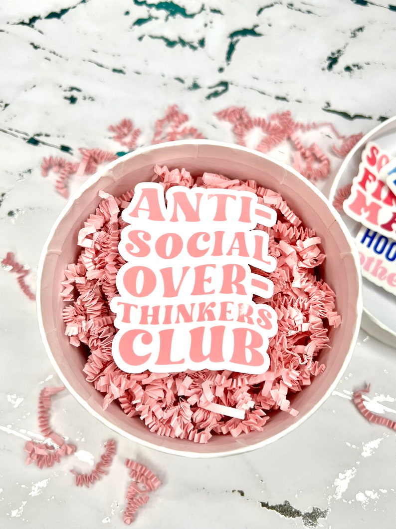 May include: A pink and white sticker with the text "Anti-Social Over-Thinkers Club" in a retro font style. The sticker is in a white round container filled with pink crinkle paper.