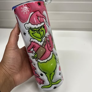 May include: A pink and green stainless steel tumbler with a Grinch Christmas design. The Grinch is wearing a Santa hat and holding a cup of coffee. The tumbler has a snowflake and stars on it.