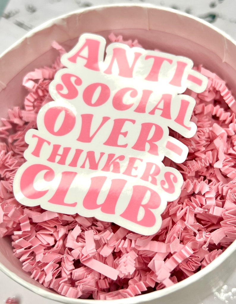 May include: A pink sticker with the text "ANTI-SOCIAL OVER-THINKERS CLUB" in white letters. The sticker is sitting on a bed of pink crinkle paper.