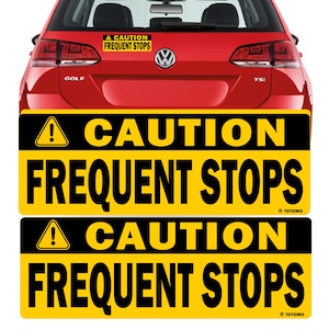 以下が含まれることがあります： 「CAUTION FREQUENT STOPS」というテキストと黒い感嘆符が書かれた黄色と黒の注意喚起サイン2つ。サインは赤い車に貼られています。
