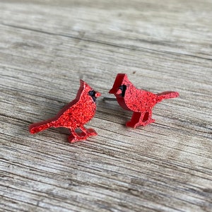 May include: A pair of red glitter cardinal earrings. The cardinals are facing to the right and have black eyes and beaks.