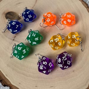 May include: A collection of colorful, polyhedral dice earrings in various colors, including blue, orange, green, yellow, and purple. Each die has white numbers and is attached to a silver earring hook. The earrings are displayed on a wooden surface.