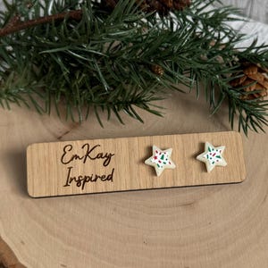 May include: A pair of white star-shaped earrings with red and green sprinkles. The earrings are displayed on a wooden board with the text "EmKay Inspired".