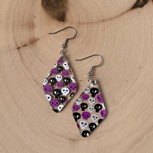 May include: A pair of diamond-shaped earrings with a clear background, adorned with a pattern of black, white, and purple skulls. The earrings have silver-colored hooks and are displayed on a wooden surface.