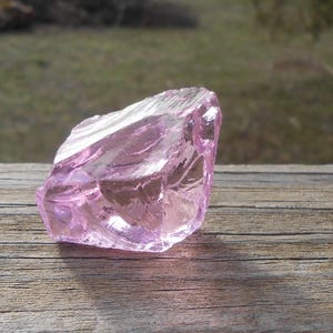 May include: A light purple, translucent rock crystal with a jagged, irregular surface. The crystal sits on a weathered wooden surface, with a blurred natural backdrop.