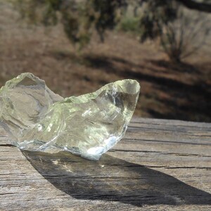 May include: A large, clear, light green crystal with a rough, irregular shape. The crystal is sitting on a wooden surface.