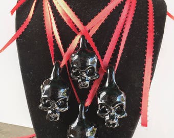 Op de afbeelding: Vier zwarte schedelhangers met rode linten, gepresenteerd op een zwarte fluwelen buste. De glanzende schedels hangen aan dunne, felrode linten. De buste staat tegen een witte achtergrond. Gothic sieraden.