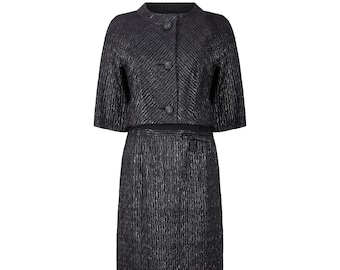 May include: A black and silver two-piece suit with a short-sleeved jacket and a skirt. The jacket has a round neckline and three buttons down the front. The skirt is knee-length and has a straight silhouette.