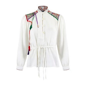 May include: A white long-sleeved shirt with a mandarin collar and a tie belt. The shirt has colourful embroidered details on the shoulders and a tassel hanging from the collar.