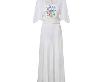 May include: A white dress with a square neckline and short sleeves. The bodice features a floral embroidery in pink, blue, and green. A crocheted belt cinches the waist, and the skirt flows to the floor. The dress has a vintage or bohemian style.