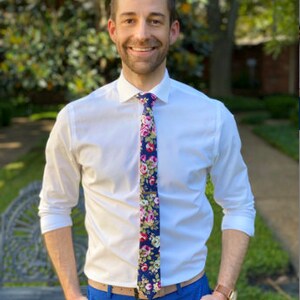 May include: A man wearing a white button-down shirt with a floral patterned tie. He is wearing blue pants and has a brown belt. He is standing in front of a green bush.