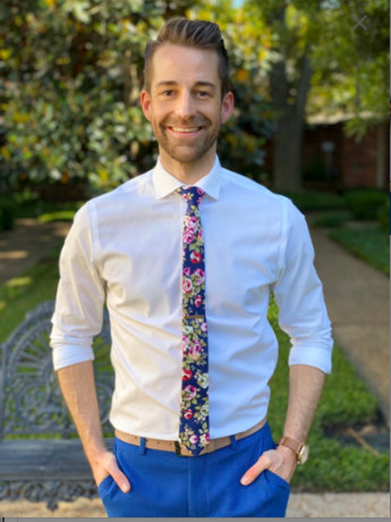 May include: A man wearing a white button-down shirt with a floral patterned tie. He is wearing blue pants and has a brown belt. He is standing in front of a green bush.