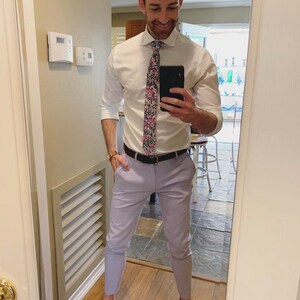 May include: A man wearing a white button-down shirt with a floral patterned tie, lavender pants, and brown leather shoes. He is standing in front of a mirror and taking a selfie.