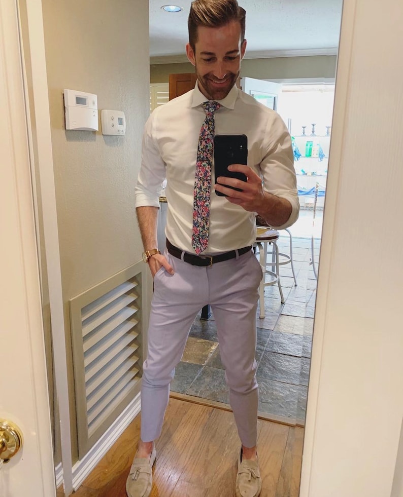 May include: A man wearing a white button-down shirt with a floral patterned tie, lavender pants, and brown leather shoes. He is standing in front of a mirror and taking a selfie.