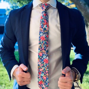 May include: A navy blue blazer with a white dress shirt and a floral patterned tie. The tie has a mix of pink, blue, and green flowers on a dark blue background.