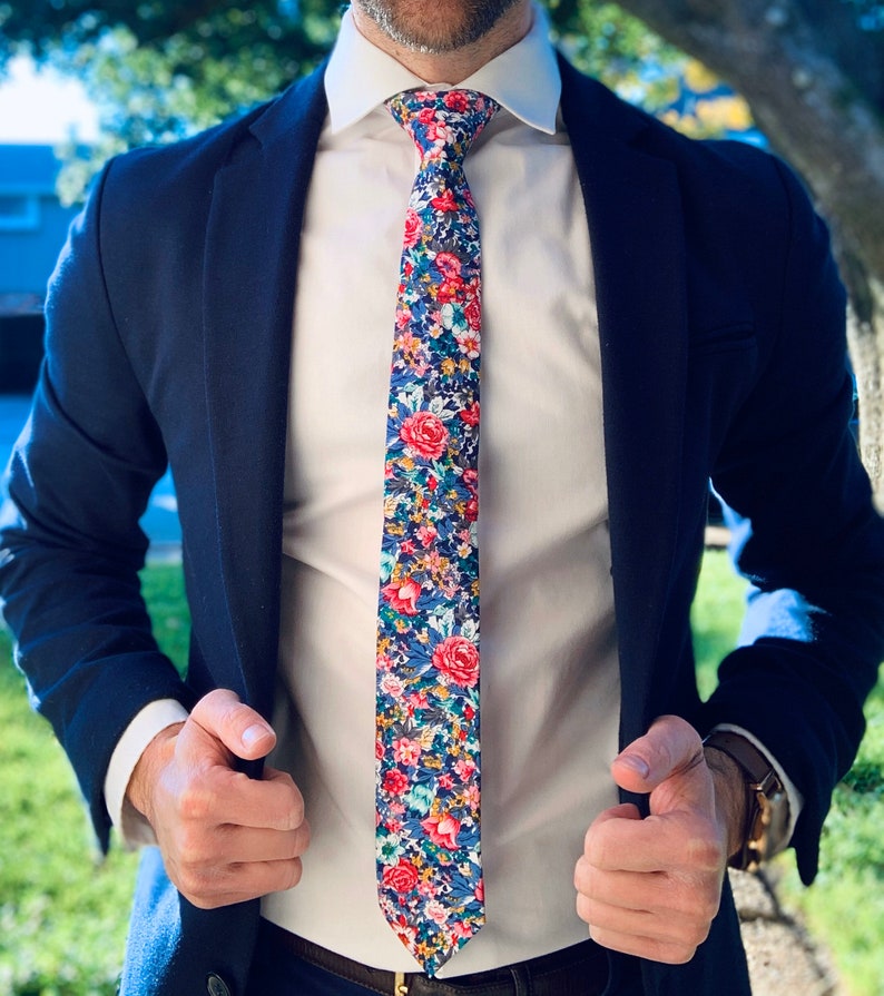 May include: A navy blue blazer with a white dress shirt and a floral patterned tie. The tie has a mix of pink, blue, and green flowers on a dark blue background.