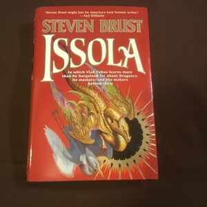 May include: A red paperback book with a gold sunburst design on the cover. The book is titled "Issola" and features the author's name, Steven Brust, in large, gold letters. The book's blurb reads "In which Vlad Taltos learns more than he bargained for about Dragaera, its masters, and the makers behind them."