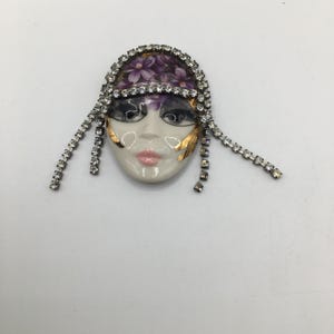 May include: A ceramic face brooch featuring a white complexion, pink lips, and dark eyes. The forehead is adorned with a purple floral pattern, while gold accents decorate the cheeks. The piece is framed by clear rhinestone chains on both sides.