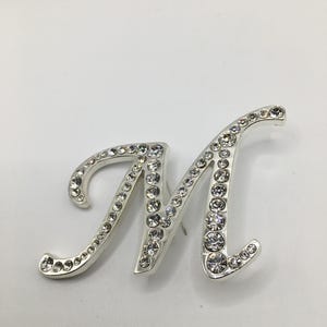 May include: Silver letter "M" embellished with numerous clear, sparkling rhinestones. The cursive-style initial is set against a white background. The rhinestones are of varying sizes, adding dimension to the letter.