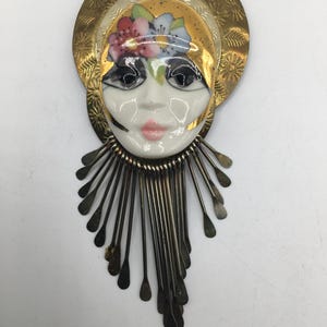 May include: A decorative brooch featuring a central porcelain face with painted features, pink lips, and a floral headpiece. The face is framed by a circular brass halo with etched sunburst patterns and a fringe of dangling silver-toned metal rods.