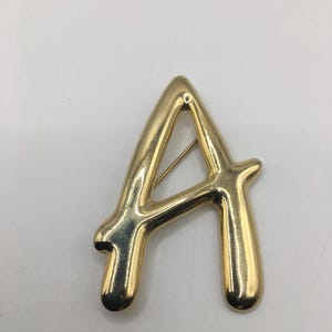 May include: A gold-colored letter "A" brooch. The brooch is in a cursive style with a pin on the back for attachment. The letter is a solid, three-dimensional design, with a shiny, reflective surface. The brooch is set against a white background.