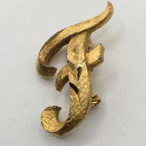 May include: A gold-toned metal brooch in the shape of the letter "F". The brooch has a textured surface and a pin closure on the back. The design features a stylized, cursive font with a decorative flourish.