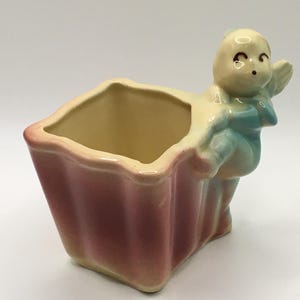 May include: A vintage ceramic planter in a rectangular shape with a scalloped rim. The planter is a gradient of pink to yellow. A small cherub figure in light blue and cream is attached to the side.