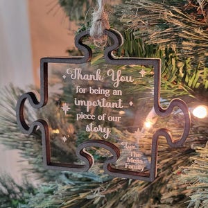 Peut inclure: Une décoration de Noël en forme de pièce de puzzle en bois brun avec un insert en acrylique transparent. La décoration porte le texte "Thank You for being an important piece of our story" et "Love The Mejia Family" gravé dessus.