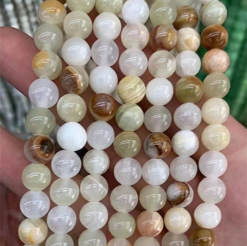 Puede incluir: Hilo de cuentas redondas de piedras preciosas en tonos blanco, crema y marr&oacute;n. Las cuentas son lisas y pulidas, con variaciones de color y patr&oacute;n. Las cuentas est&aacute;n ensartadas, listas para la fabricaci&oacute;n de joyas.