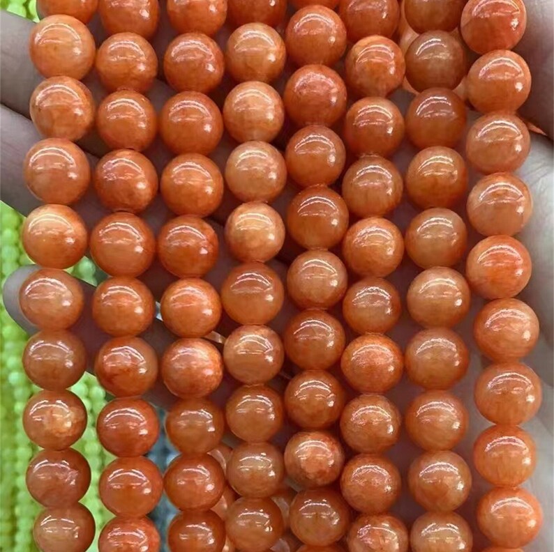 Puede incluir: Primer plano de un hilo de cuentas redondas de piedras preciosas de color naranja. Las cuentas tienen un acabado pulido y brillante y est&aacute;n ensartadas, mostrando su forma esf&eacute;rica y variaciones de color. Las cuentas miden aproximadamente 8 mm de di&aacute;metro.