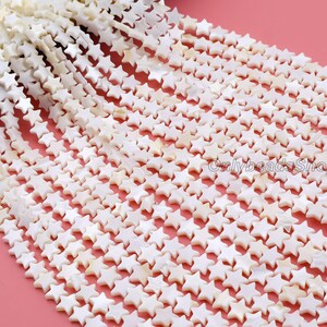 May include: Close-up of multiple strands of white, star-shaped beads, possibly for jewellery. The beads have a pearlescent finish and are set against a pink backdrop. The text "Onlybeads Shop" is visible.