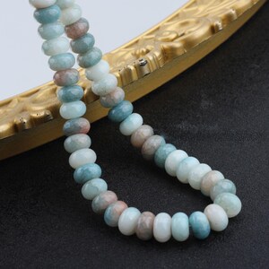 Pu&ograve; includere: Una collana di perline di pietre preziose nei toni del blu, bianco e marrone. Le perline rotonde sono infilate insieme. Le perline sono esposte su una superficie color oro su sfondo nero.